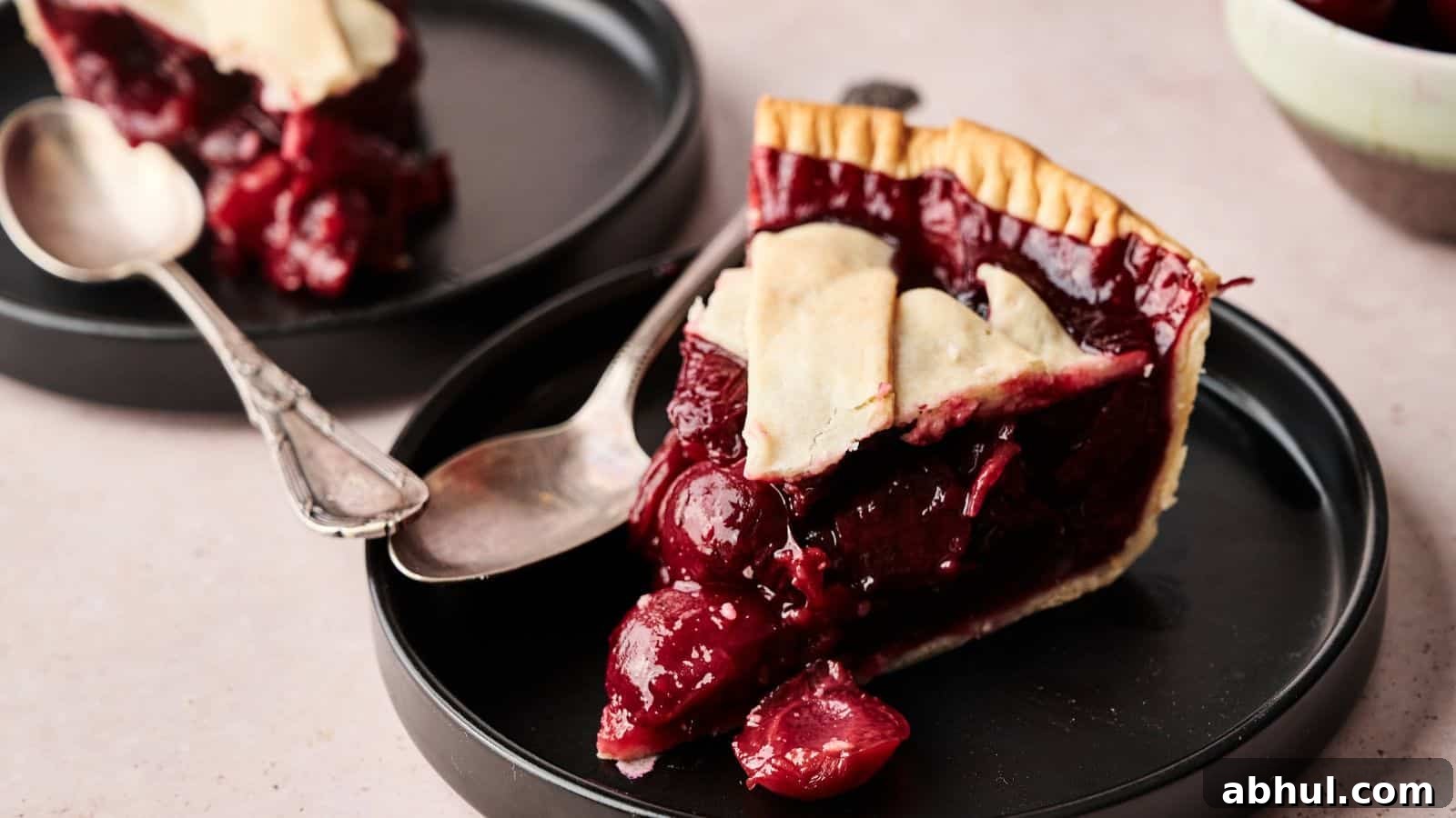 A slice of cherry pie with a flaky crust is served on a black plate with a spoon; another plate with pie is in the background.
