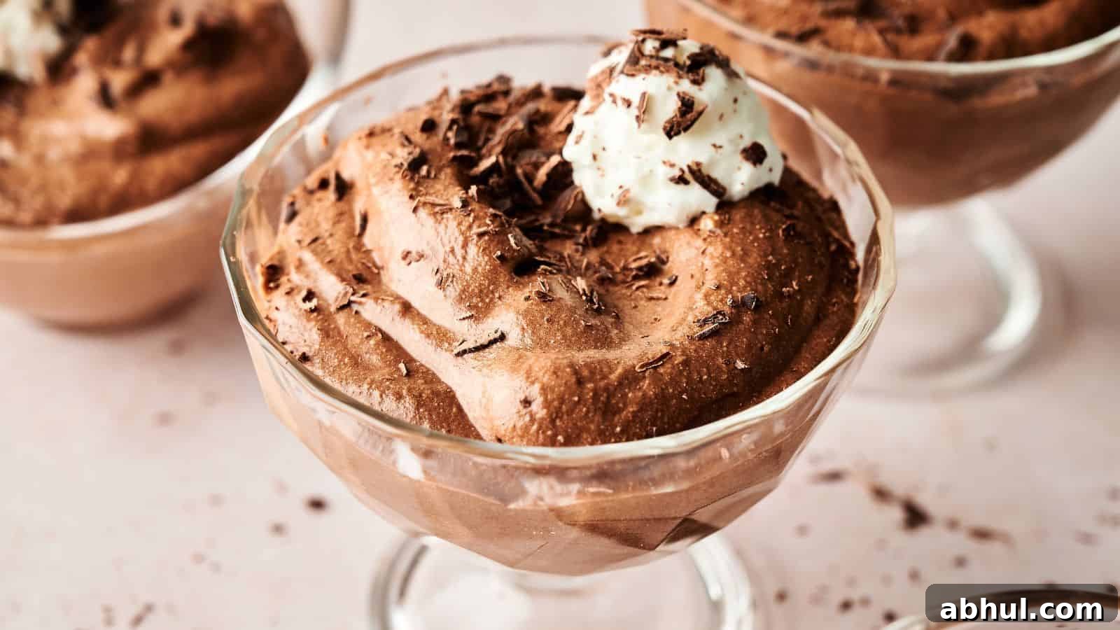 A glass dessert cup filled with chocolate mousse, topped with whipped cream and chocolate shavings, placed on a light-colored surface.