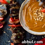 Flank steak, red onions and red bell peppers on wooden skewers with a bowl of peanut sauce next to them.