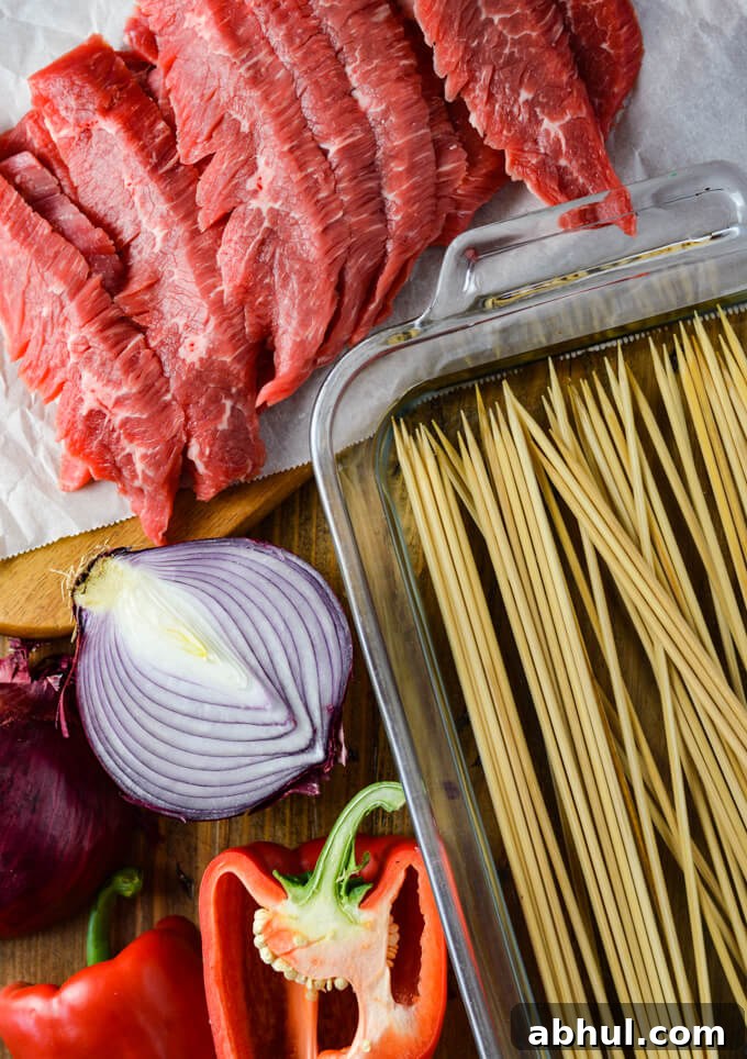 Strips of raw flank steak with wooden skewers in a glassware pan of water. Halved red onion and red bell pepper sit next to it.