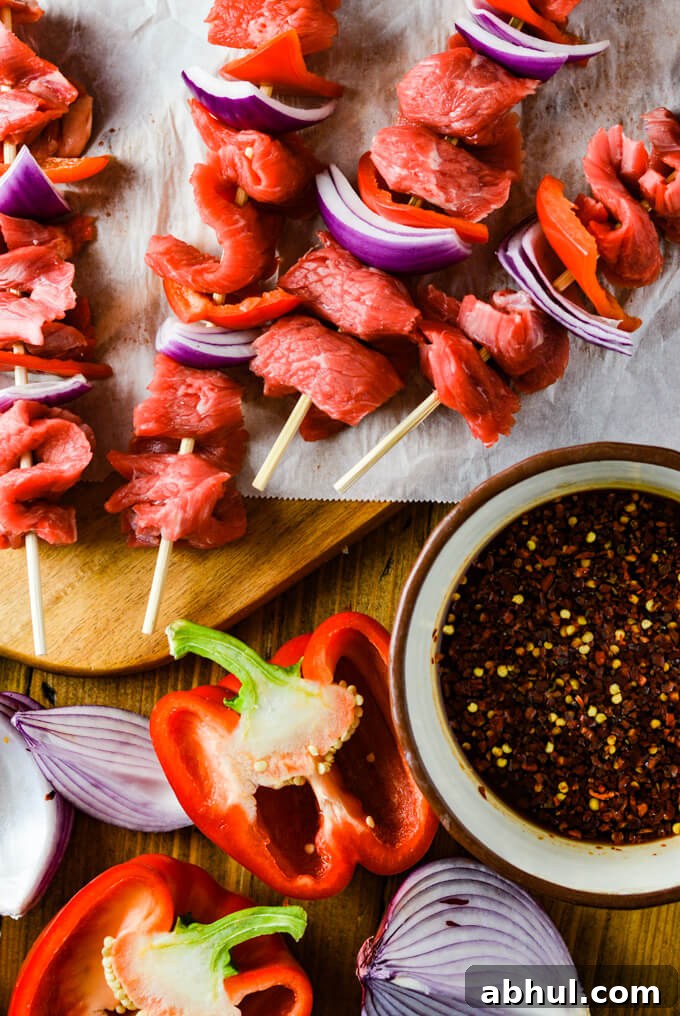Raw flank stead, red onions and red bell peppers on four wooden skewers laying on a wooden cutting board with parchment paper. A bowl of marinade and halved red onions and red bell peppers sit next to it.