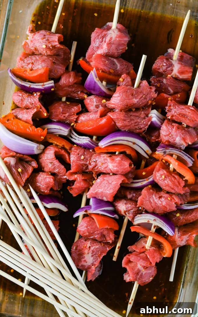 Flank steak, red onion and red bell peppers on wooden skewers sit in a glassware pan with marinade.