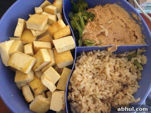 A divided dish containing cubed tofu, peanut sauce, and brown rice.