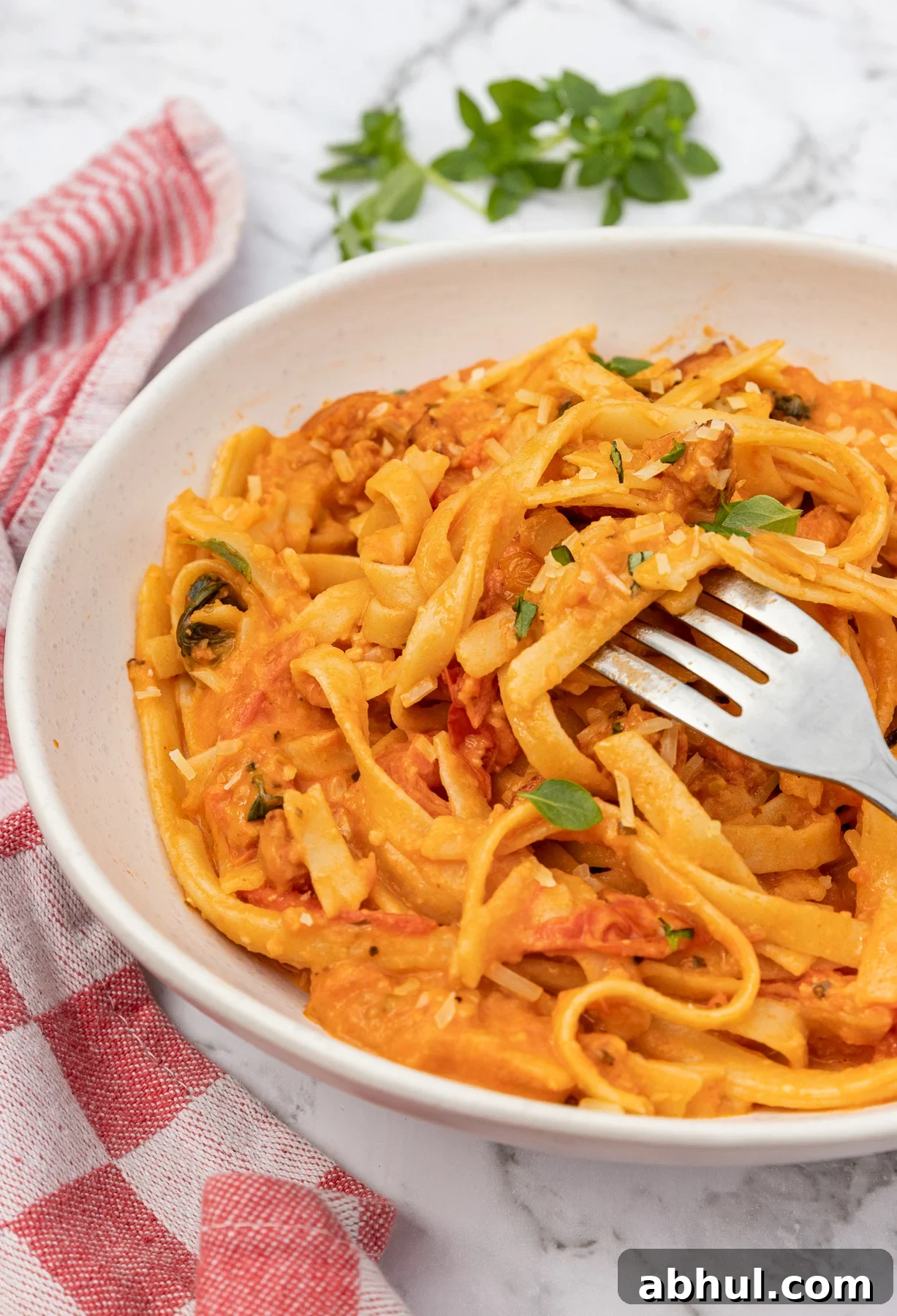 A white dish of chorizo pasta with a fork in it with a red and white checked tea towel and on a marble background.