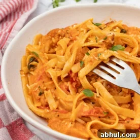 A white dish of creamy chorizo pasta with a fork in it with a red and white checked tea towel and on a marble background.