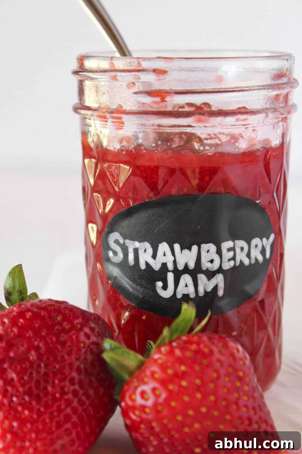 A jar of homemade strawberry jam with a spoon in the jar and fresh strawberries.