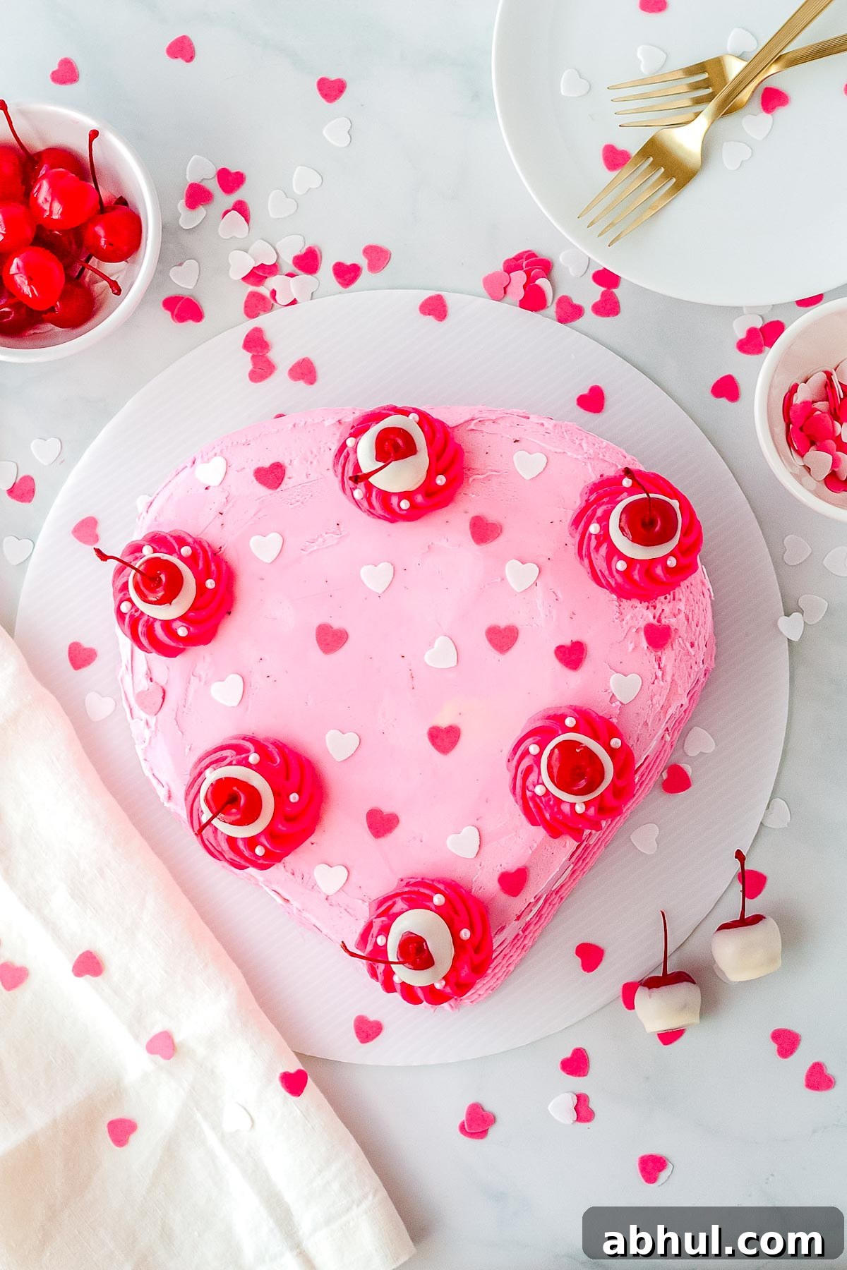 A heart cake covered in pink buttercream, heart-shaped sprinkles, and white chocolate covered cherries. 