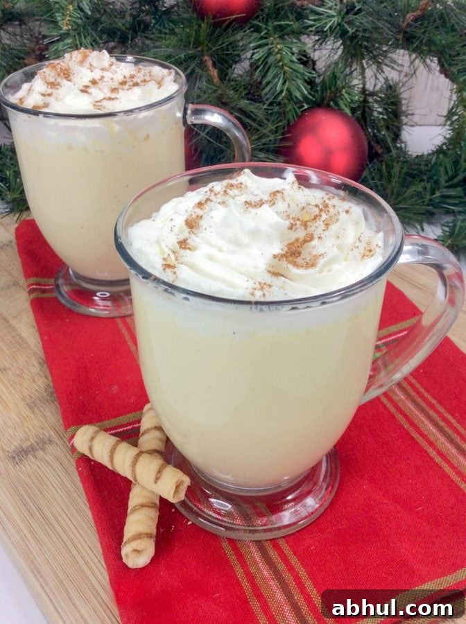 2 glass of eggnog topped with whipped cream and nutmeg on a red tablecloth.