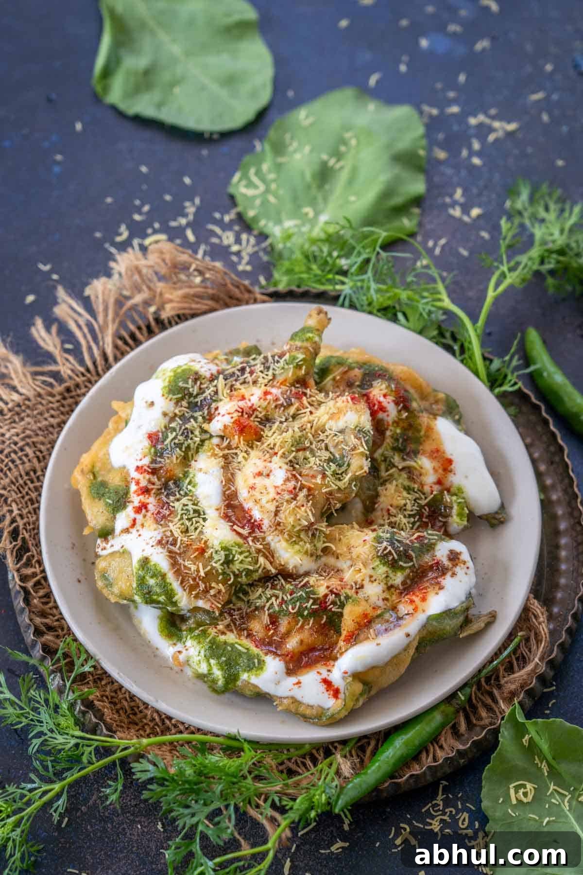 A plate of palak patta chaat topped with yogurt, sev, and spices, garnished with fresh cilantro, kept on a rustic mat with green leaves and chilies, all set against a dark background.