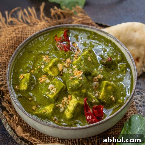 A bowl of Palak Paneer garnished with dried red chilies sits on a rustic mat, surrounded by naan bread and fresh spinach leaves for an inviting, authentic spread.