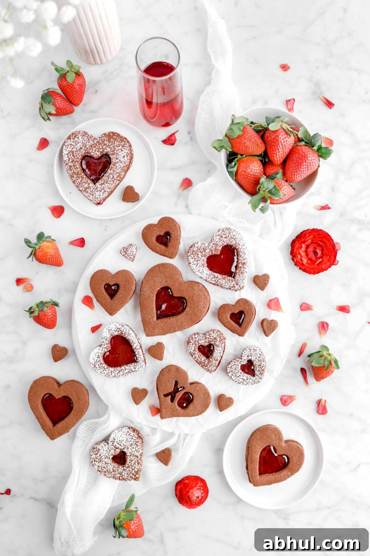 Chocolate-Strawberry Linzer Cookie Recipe for Perfect Sandwich Cookies 6 overhead shot of cookies on upside down white plate with red petals, red flowers, strawberries, and glass of red wine