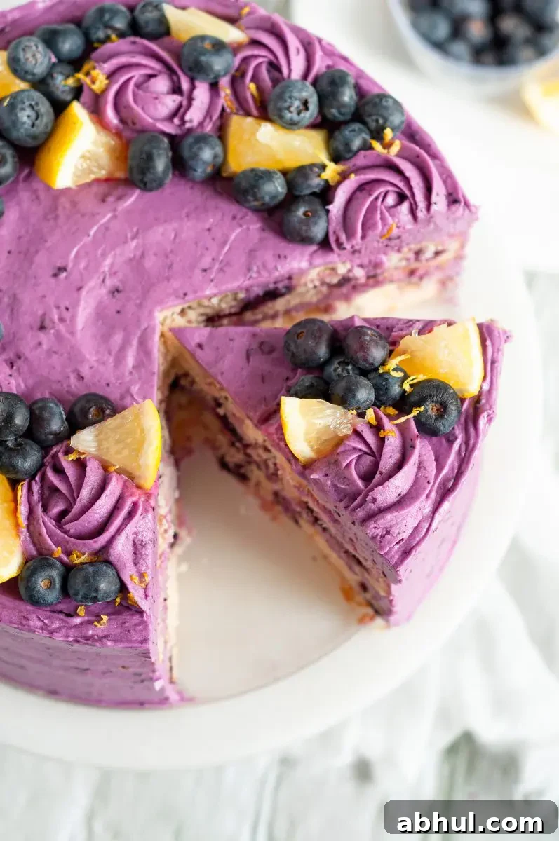 A close up picture of a purple frosted lemon blueberry layer cake. There is a large slice cut into the cake and slightly taken out of it. There are blueberries and lemon slices on the top of the cake.