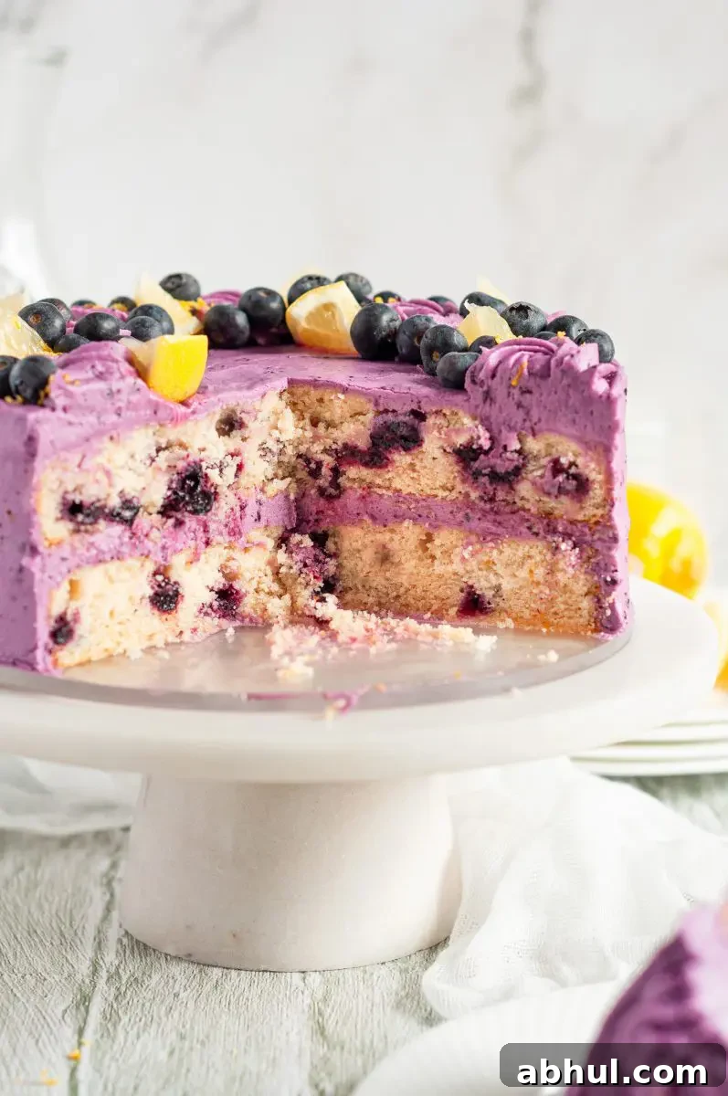 A lemon blueberry layer cake covered in purple blueberry frosting. It's sitting on a white cake stand and there are a few pieces missing allowing you to see the two cake layers inside which are speckled with blueberries.