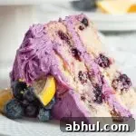 A slice of purple frosted lemon blueberry cake. It has two white cake layers full of blueberries and is topped with lemon sliced and fresh blueberries.