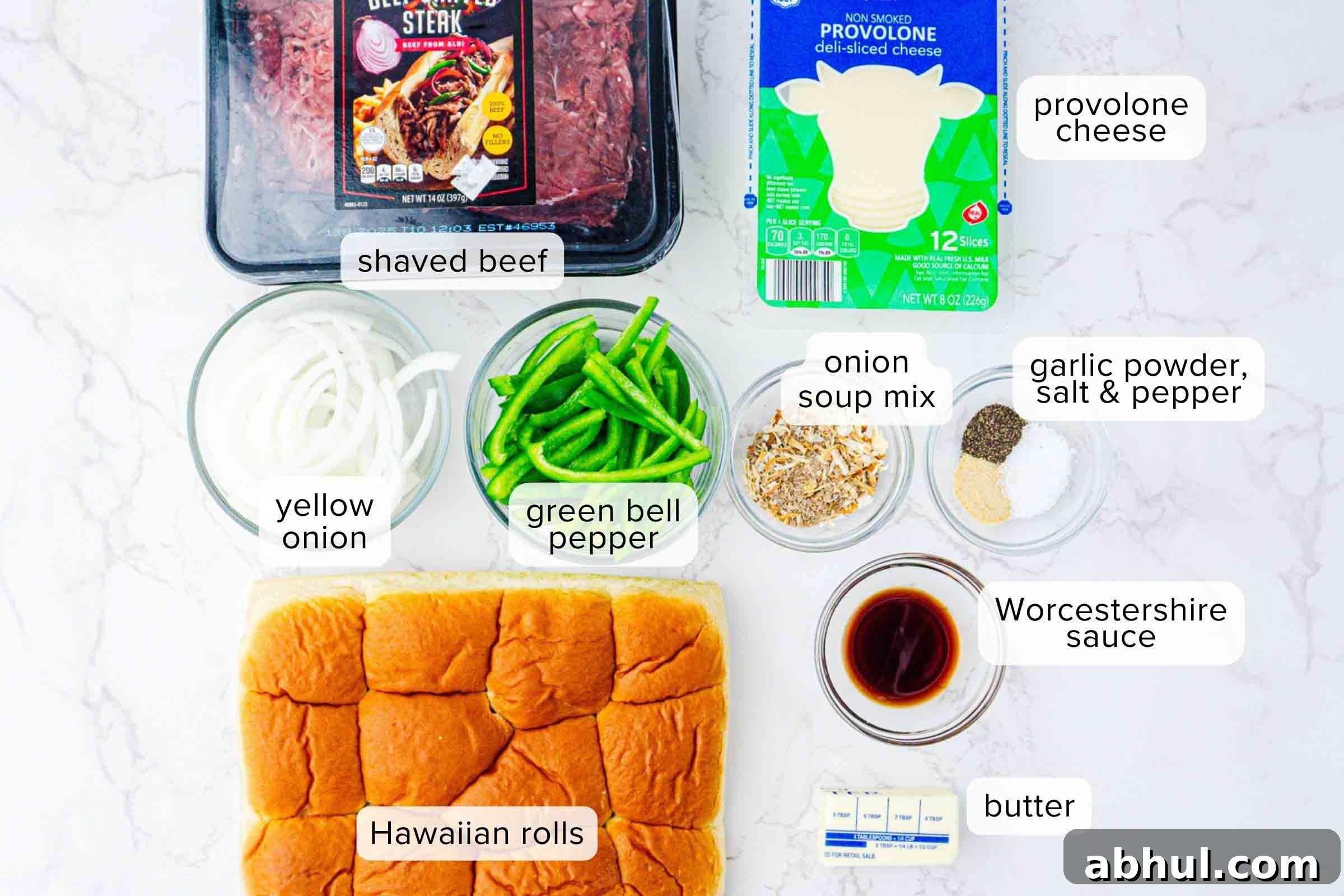 Labeled ingredients on a counter: shaved beef, provolone cheese, sliced yellow onion, sliced green bell pepper, onion soup mix, garlic powder, salt, pepper, Worcestershire sauce, butter, and Hawaiian rolls.