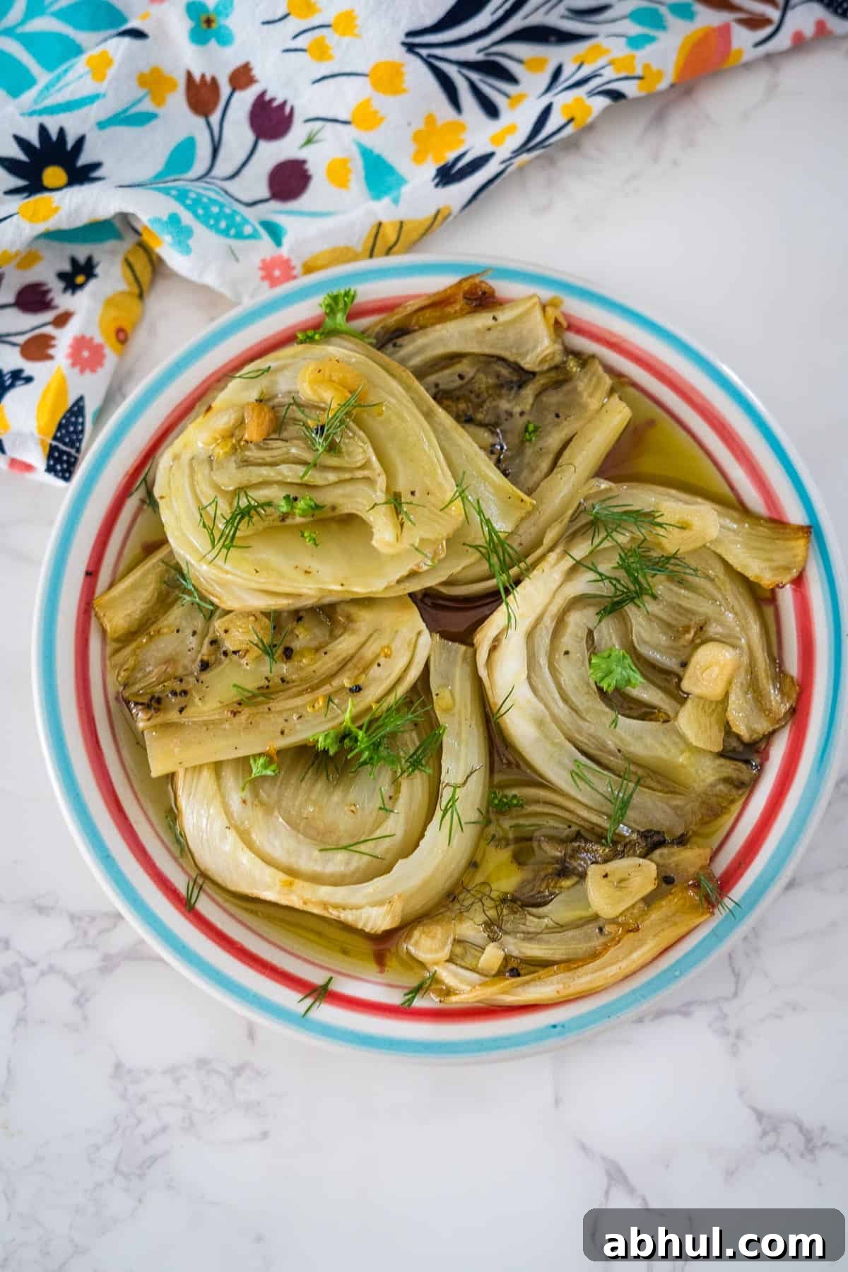 A plate of braised fennel halves garnished with herbs, pictured on a marble countertop next to a colorful cloth.