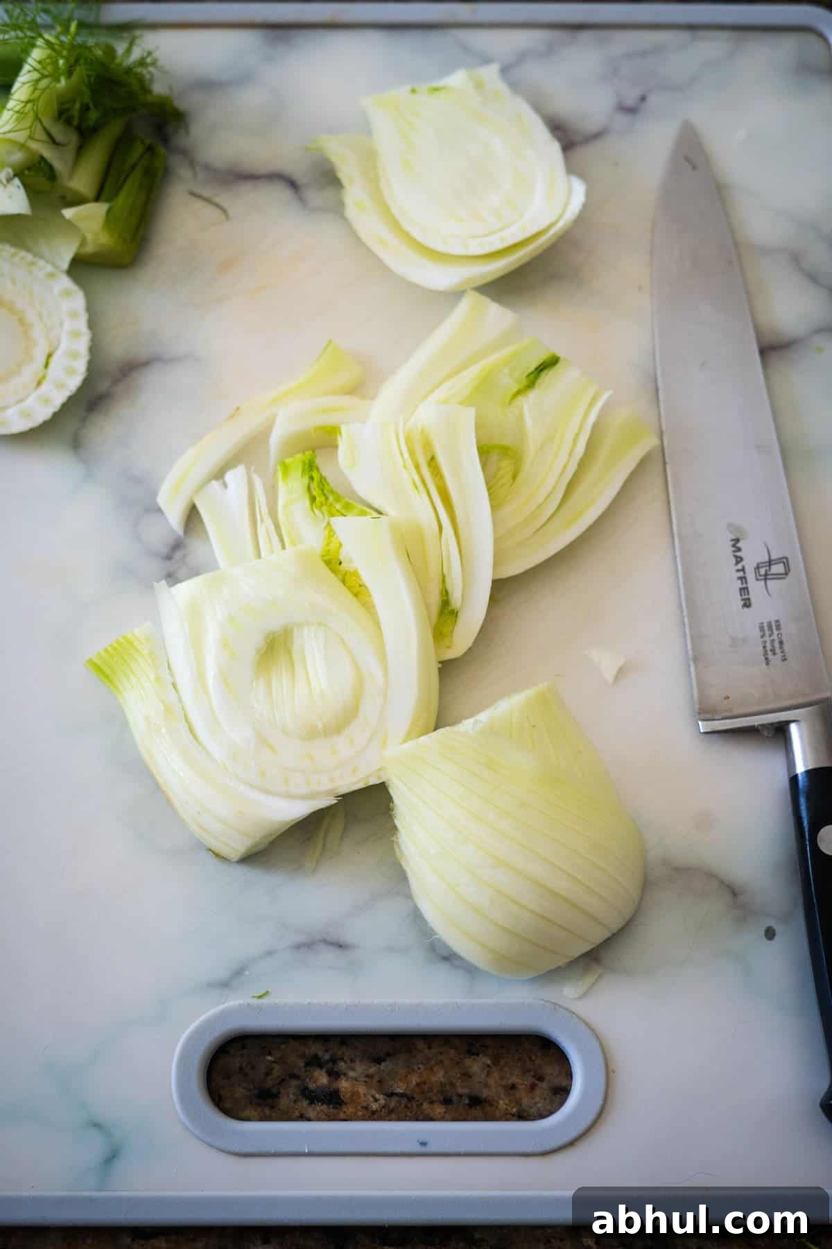 Several slices of fennel confit on a marble countertop next to a chef's knife, with a built-in countertop waste chute.