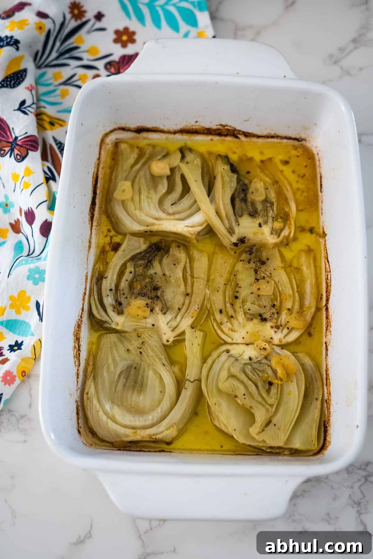 Baked fennel confit in a white ceramic dish on a marble countertop with a colorful floral napkin on the side.