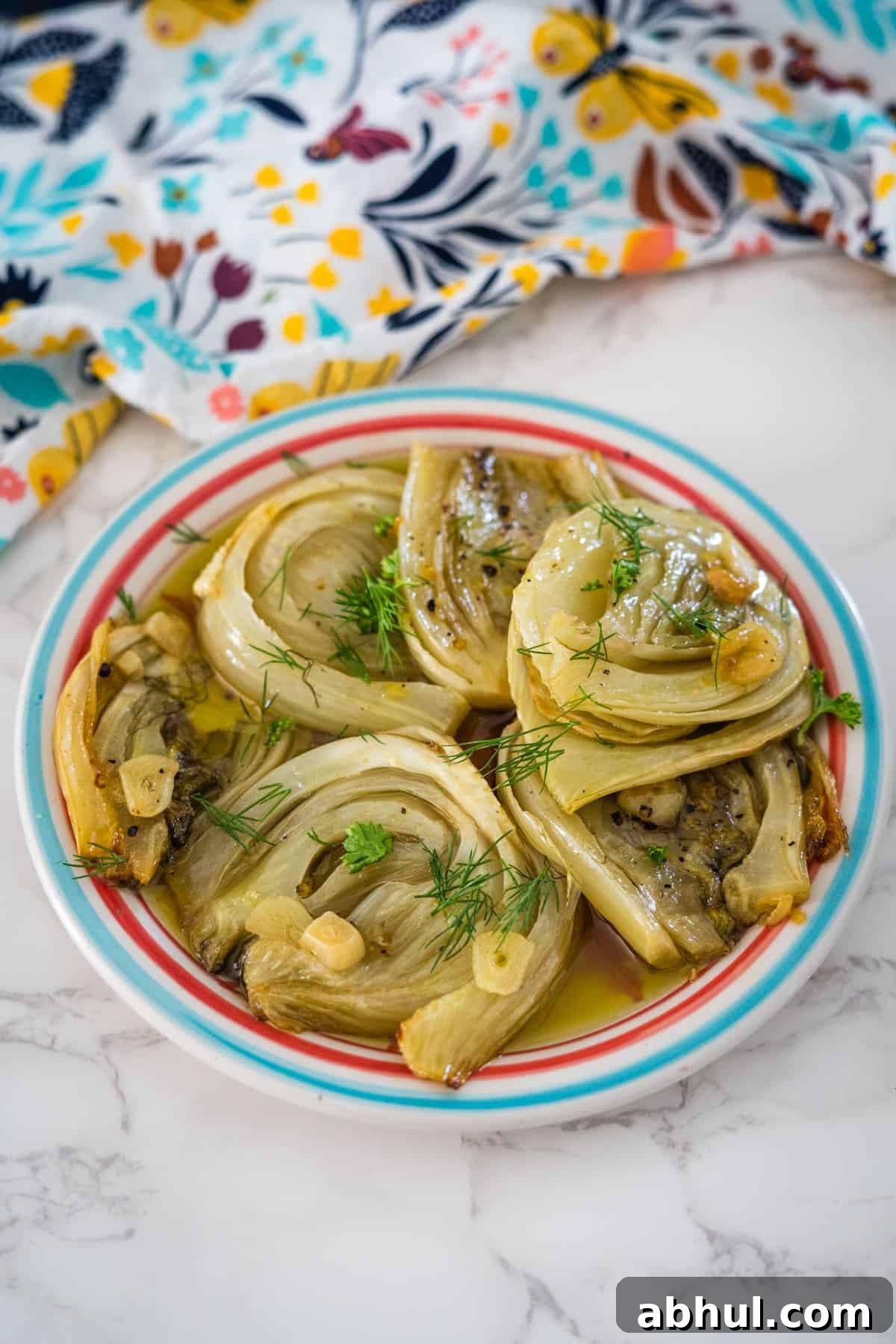 A plate of roasted fennel confit garnished with dill on a striped plate, set on a marble surface with a colorful floral cloth.