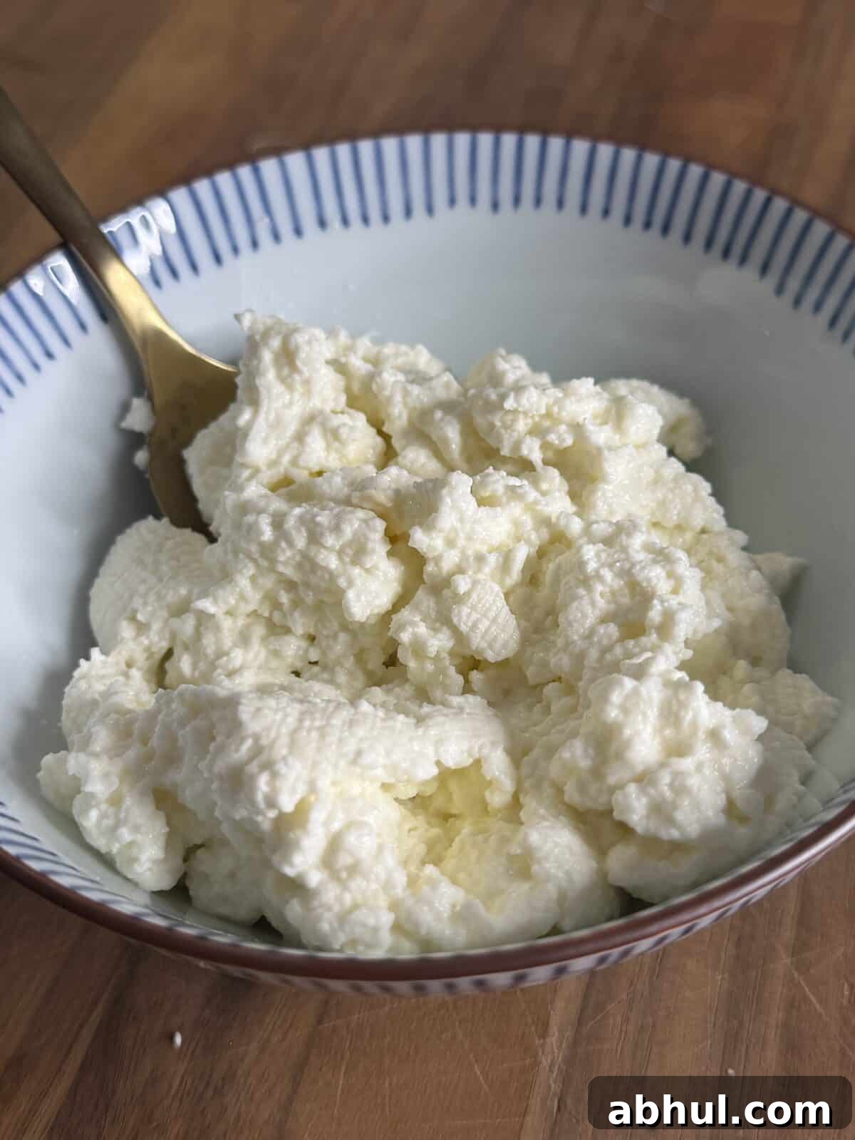 A bowl of ricotta cheese with a gold spoon on a wooden surface. The ricotta has a soft, crumbly texture and is white in color. The bowl has blue stripes along the rim.