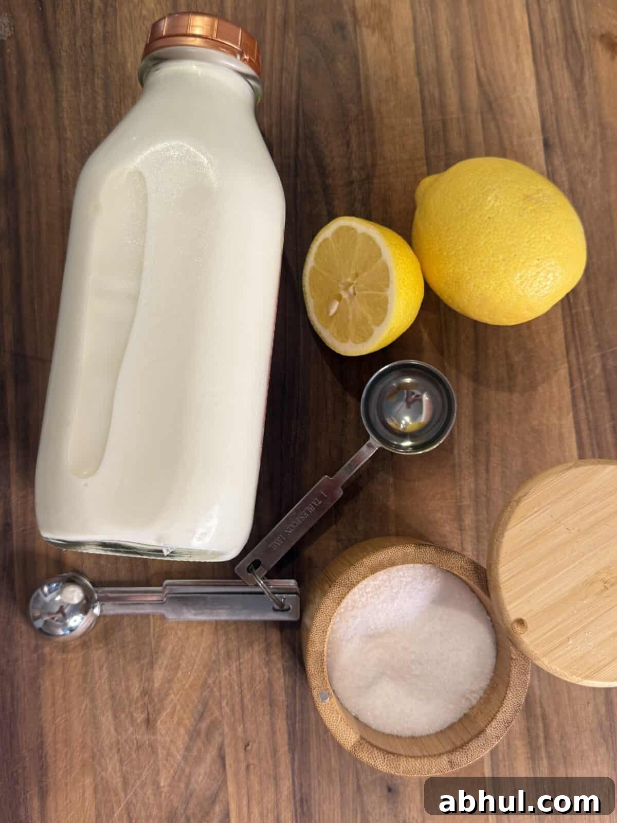 A bottle of milk, a small wooden bowl of salt, two lemons (one cut in half), and a metal measuring spoon are arranged on a wooden surface.