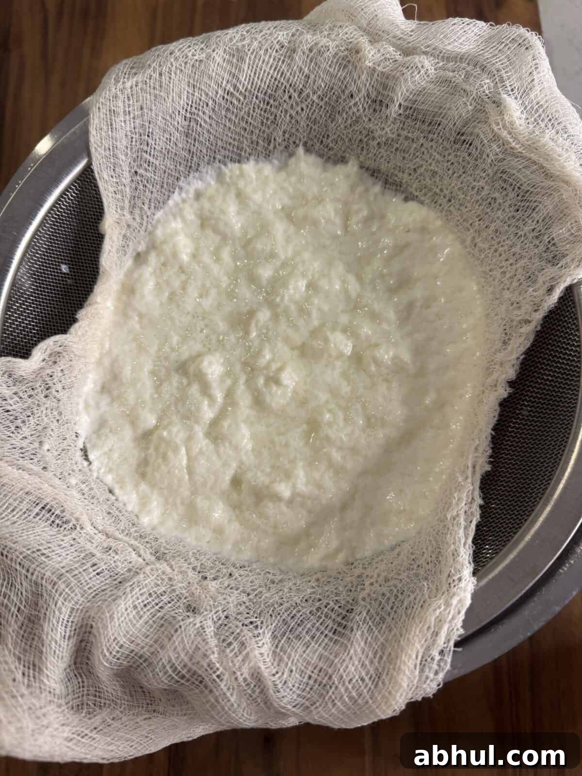 Homemade ricotta cheese curds draining in a sieve lined with cheesecloth, set over a metal bowl on a wooden surface.