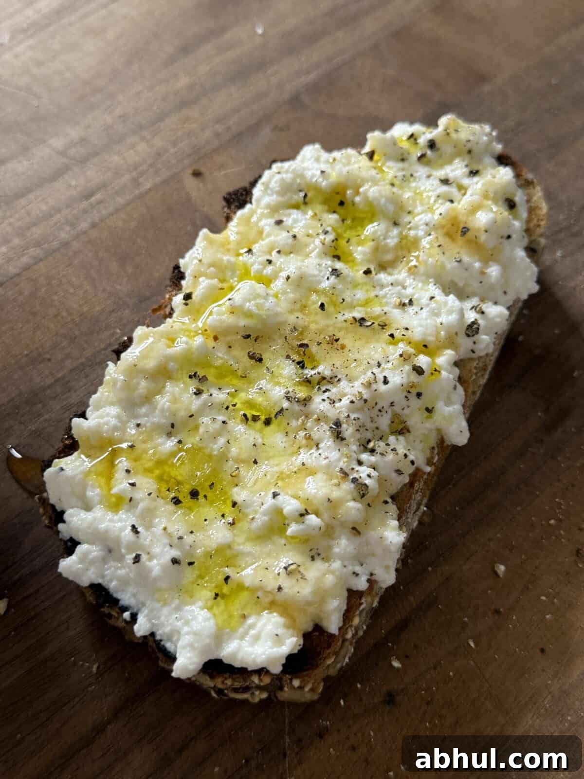 A slice of toasted bread topped with creamy ricotta cheese, a drizzle of olive oil, and freshly ground black pepper, served on a wooden surface.
