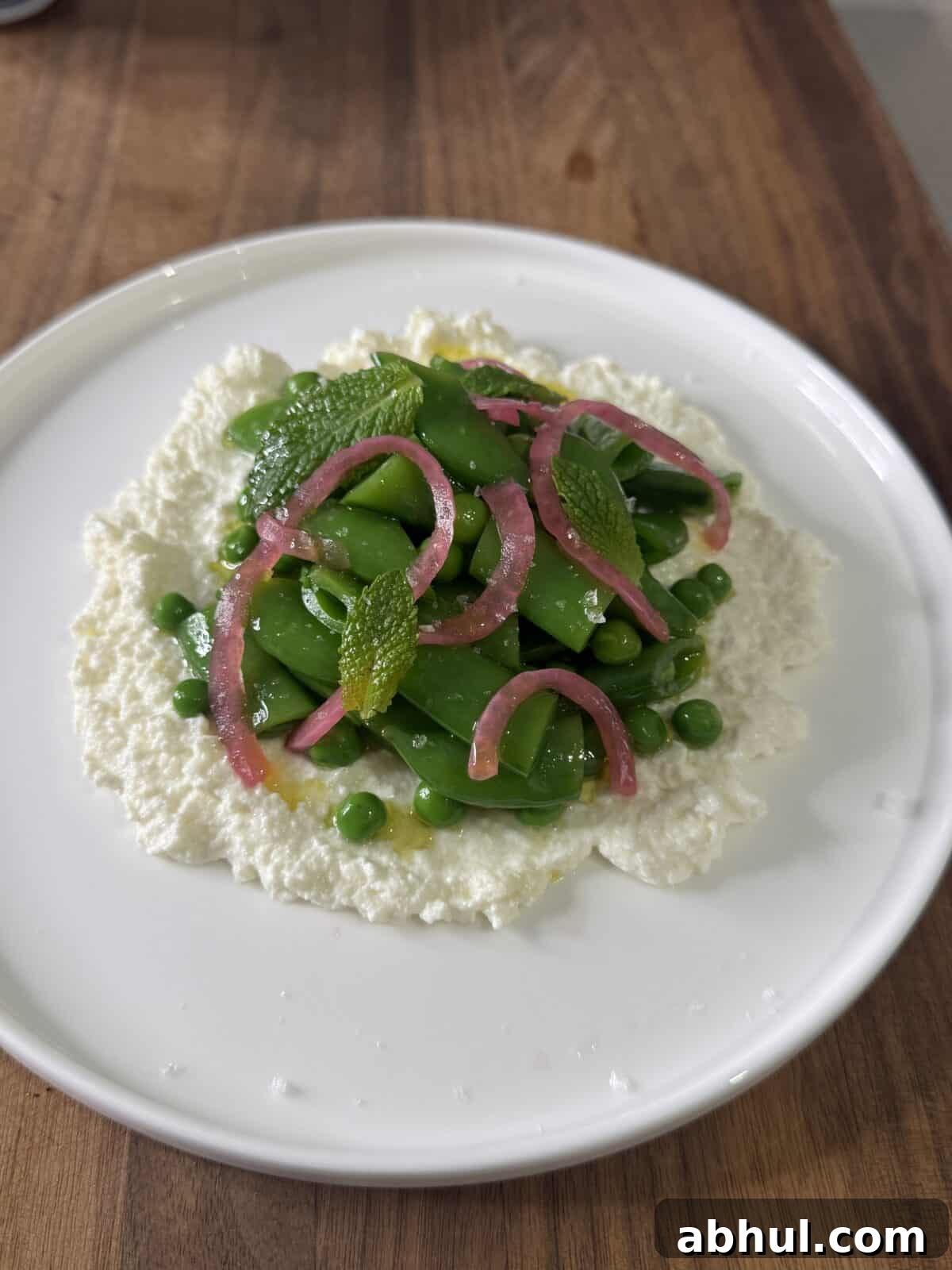 A white plate with a bed of creamy ricotta topped with green peas, sugar snap peas, pickled red onions, and fresh mint leaves, set on a wooden surface.