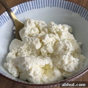 A bowl of ricotta cheese with a gold spoon on a wooden surface. The ricotta has a soft, crumbly texture and is white in color. The bowl has blue stripes along the rim.