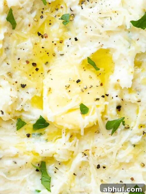 A close up of a bowl of mashed potatoes with parmesan, butter, herbs, and pepper.