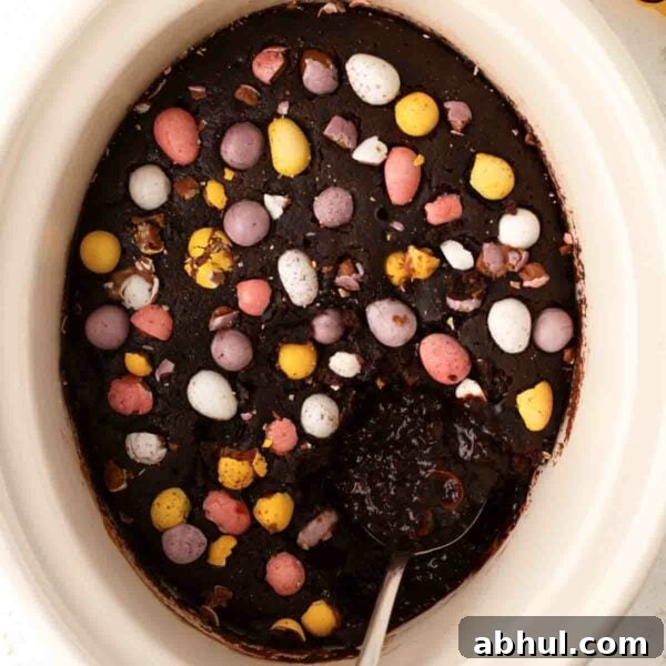 A slow cooker filled with cooked chocolate easter pudding with mini eggs on top and a spoon about to serve up.