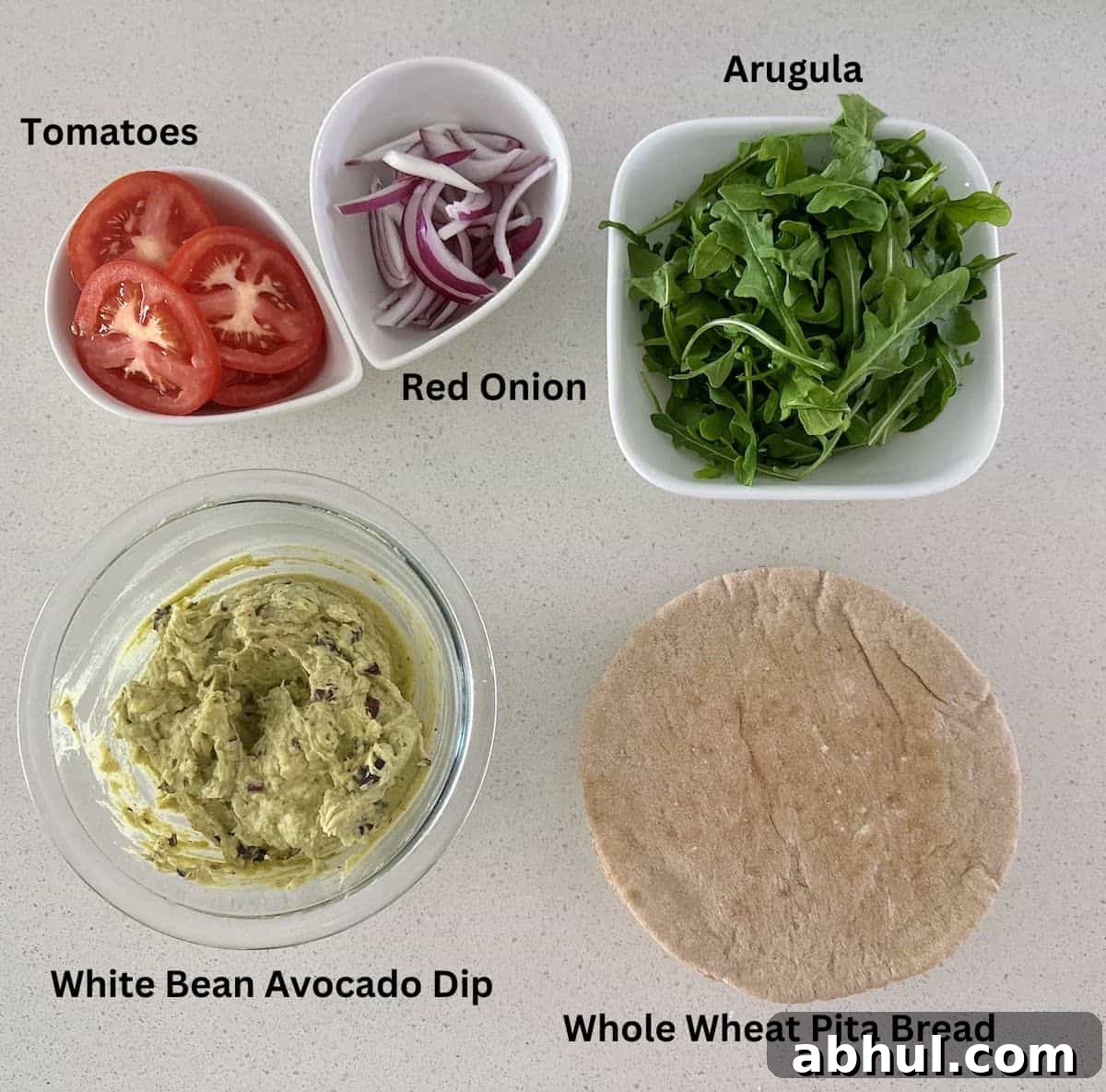 Sandwich ingredients: White bean and avocado dip, tomatoes, red onion arugula, and whole wheat pita bread.