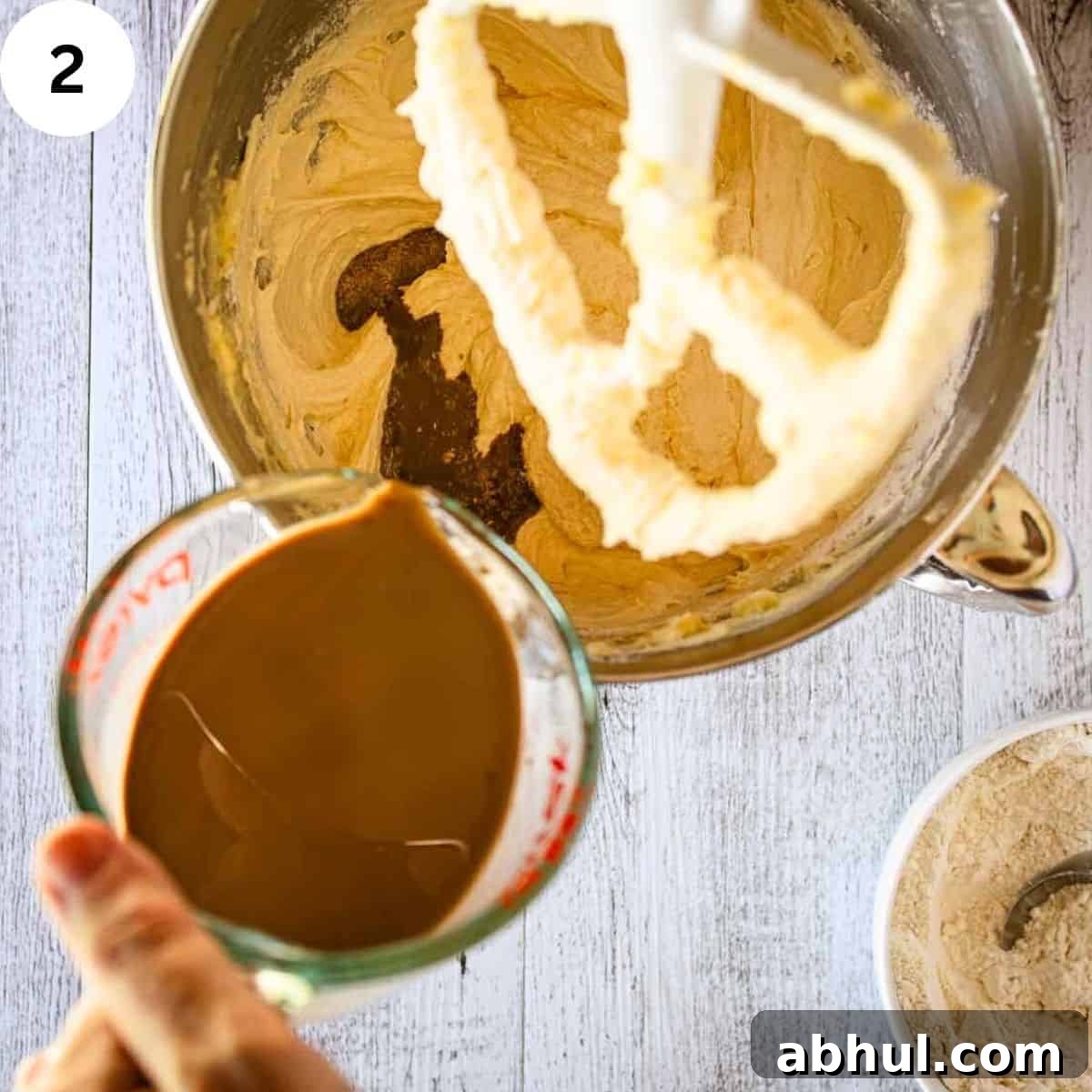 Pouring brown liquid into creamy mixture in a stand mixer bowl.