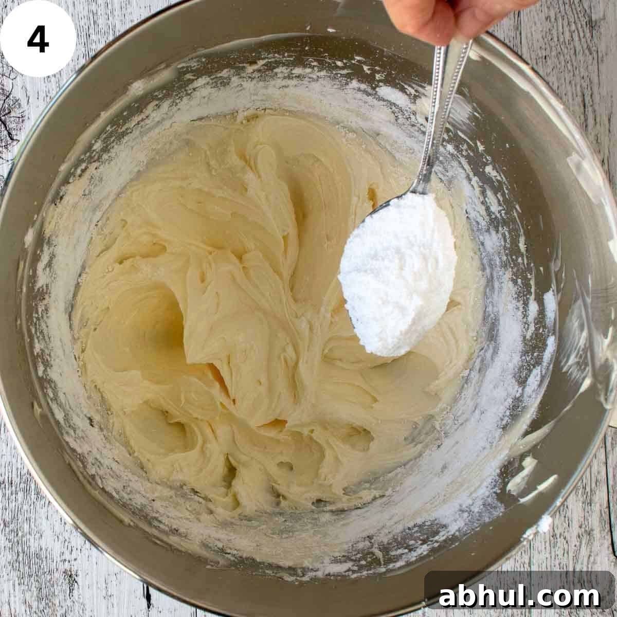 Powdered sugar being spooned into bowl containing creamy butter mixture.