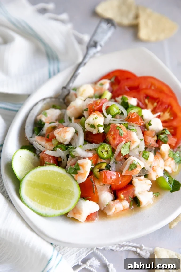 Shrimp ceviche made with onions, tomatoes, bell pepper, lime juice, and cilantro on a white serving plate with sliced tomatoes and fresh lime wedges.