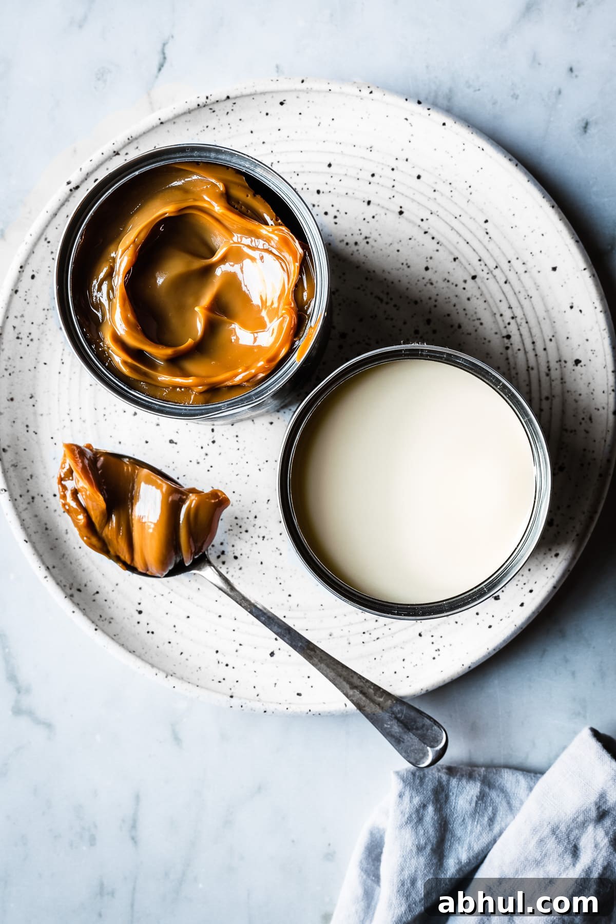 Two open cans, one of condensed milk, the other of dulce de leche, sit on a white speckled ceramic plate on a grey marble surface. A spoon holds a scoop of dulce de leche. A light blue linen napkin peeks into the bottom of the photo.