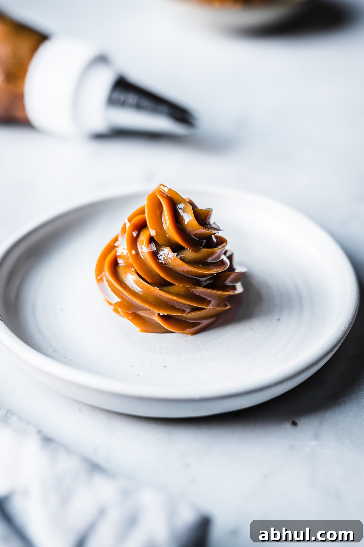 Piped dulce de leche on a white ceramic plate, sprinkled with sea salt flakes. A piping bag rests out of focus in the background. A blurry pale blue napkin peeks into the foreground.