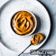A white ceramic bowl with milk caramel sauce on a white speckled ceramic plate on a grey marble surface. A spoon holds a scoop of dulce de leche. A light blue linen napkin peeks into the bottom of the photo.