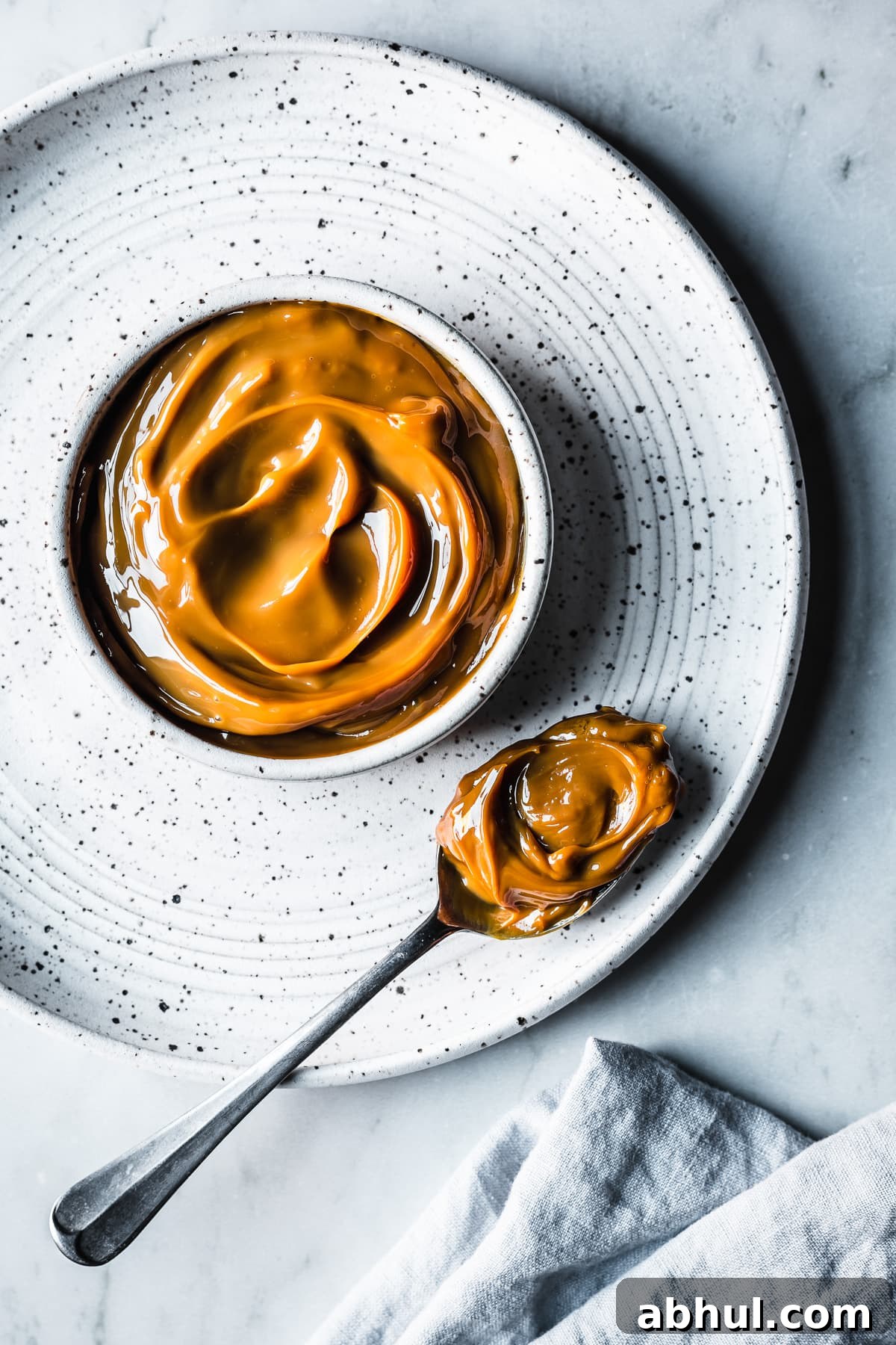 A white ceramic bowl with milk caramel sauce on a white speckled ceramic plate on a grey marble surface. A spoon holds a scoop of dulce de leche. A light blue linen napkin peeks into the bottom of the photo.
