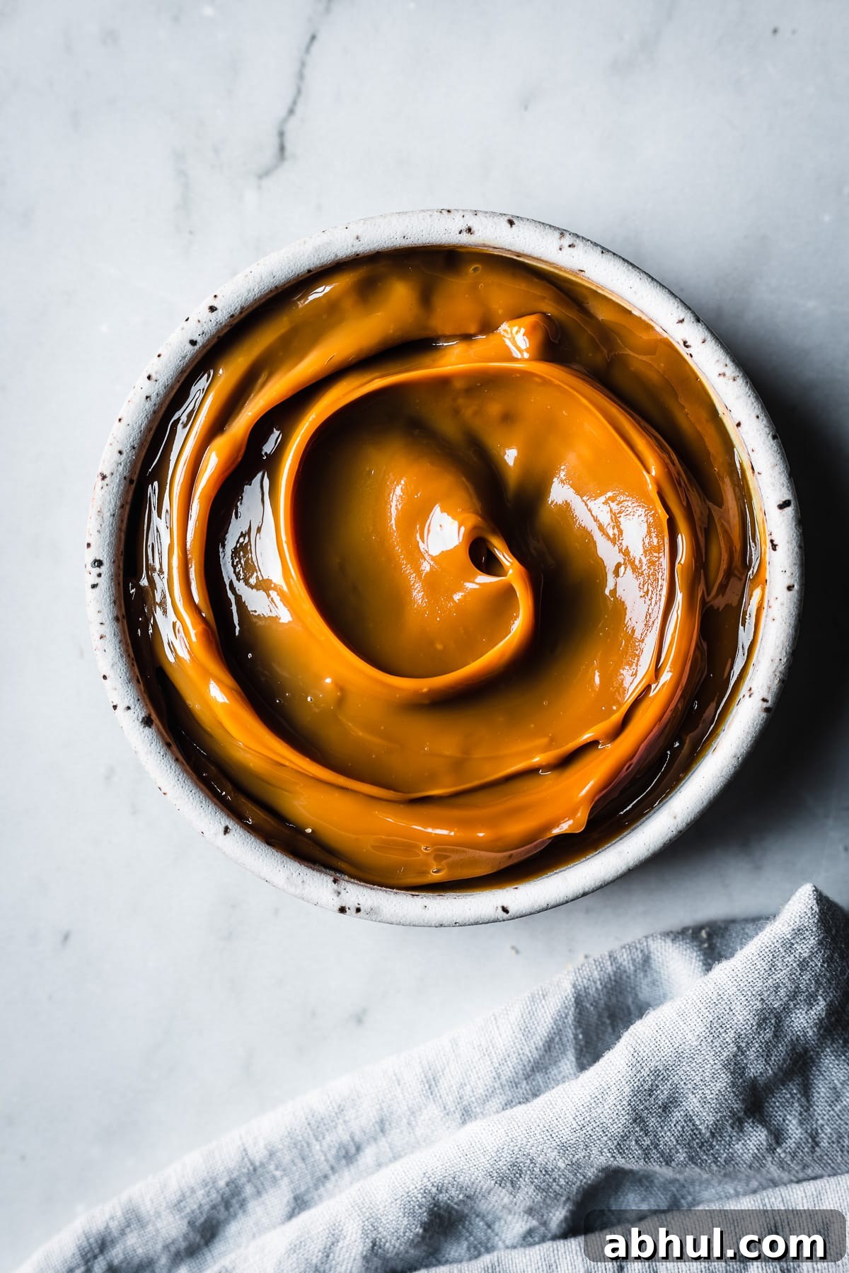 A white ceramic bowl filled with swirls of dulce de leche resting on a grey marble background. A pale blue linen napkin peeks into the bottom of the frame.