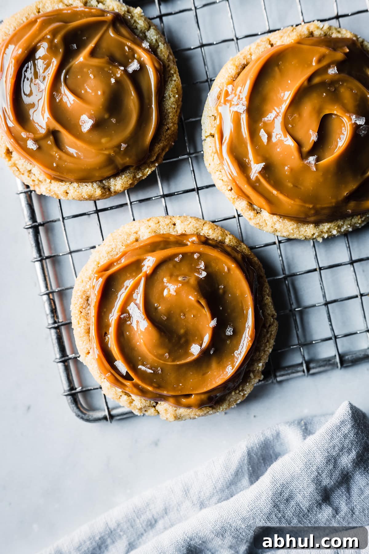 Cookies spread with milk caramel and sprinkled with sea salt on a cooling rack. The rack sits on a grey marble surface. A pale blue linen napkin peeks into the photo at bottom.