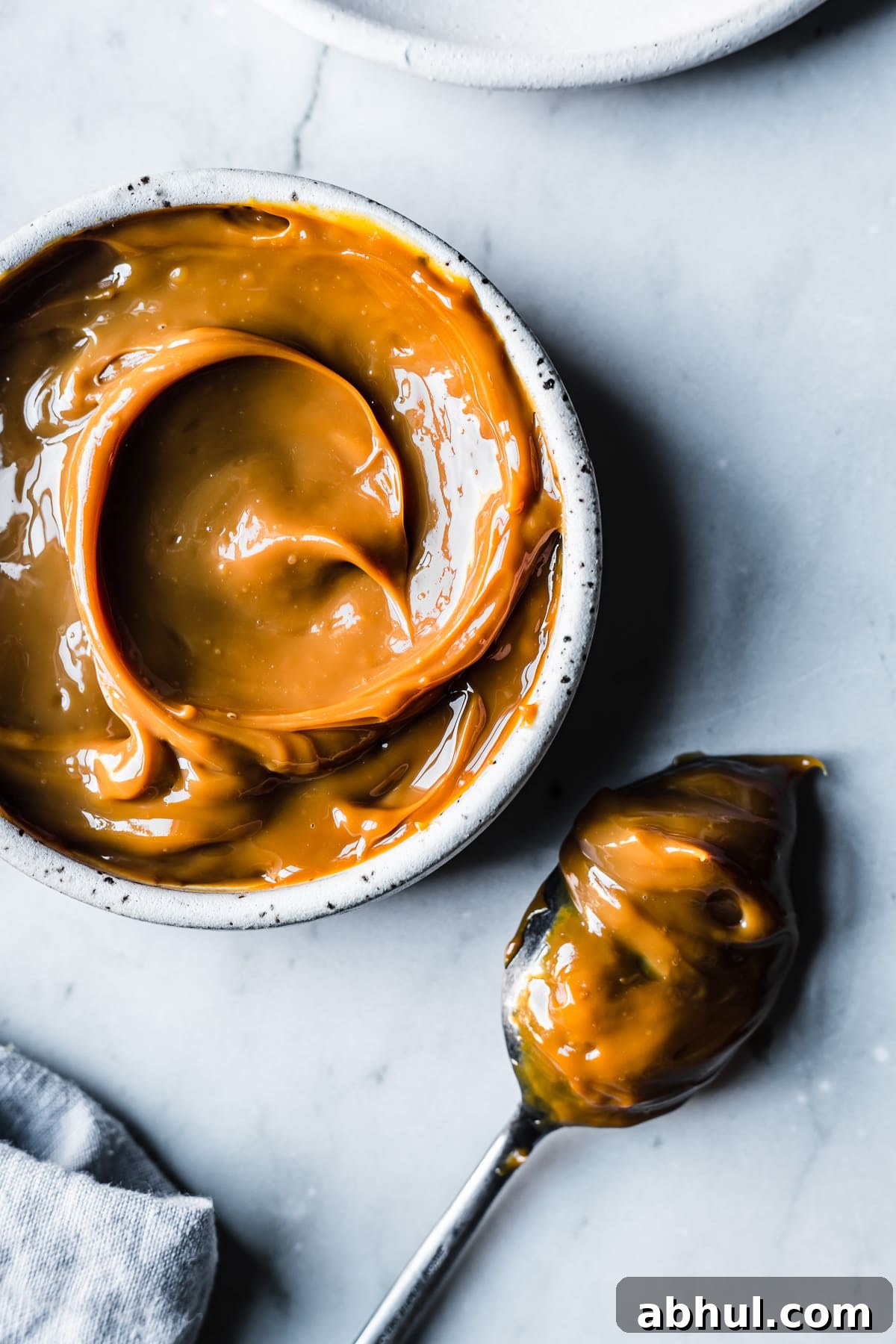 A white ceramic bowl filled with swirls of dulce de leche resting on a grey marble surface. A spoon covered in dulce de leche rests nearby. A pale blue linen napkin peeks into the bottom of the frame.