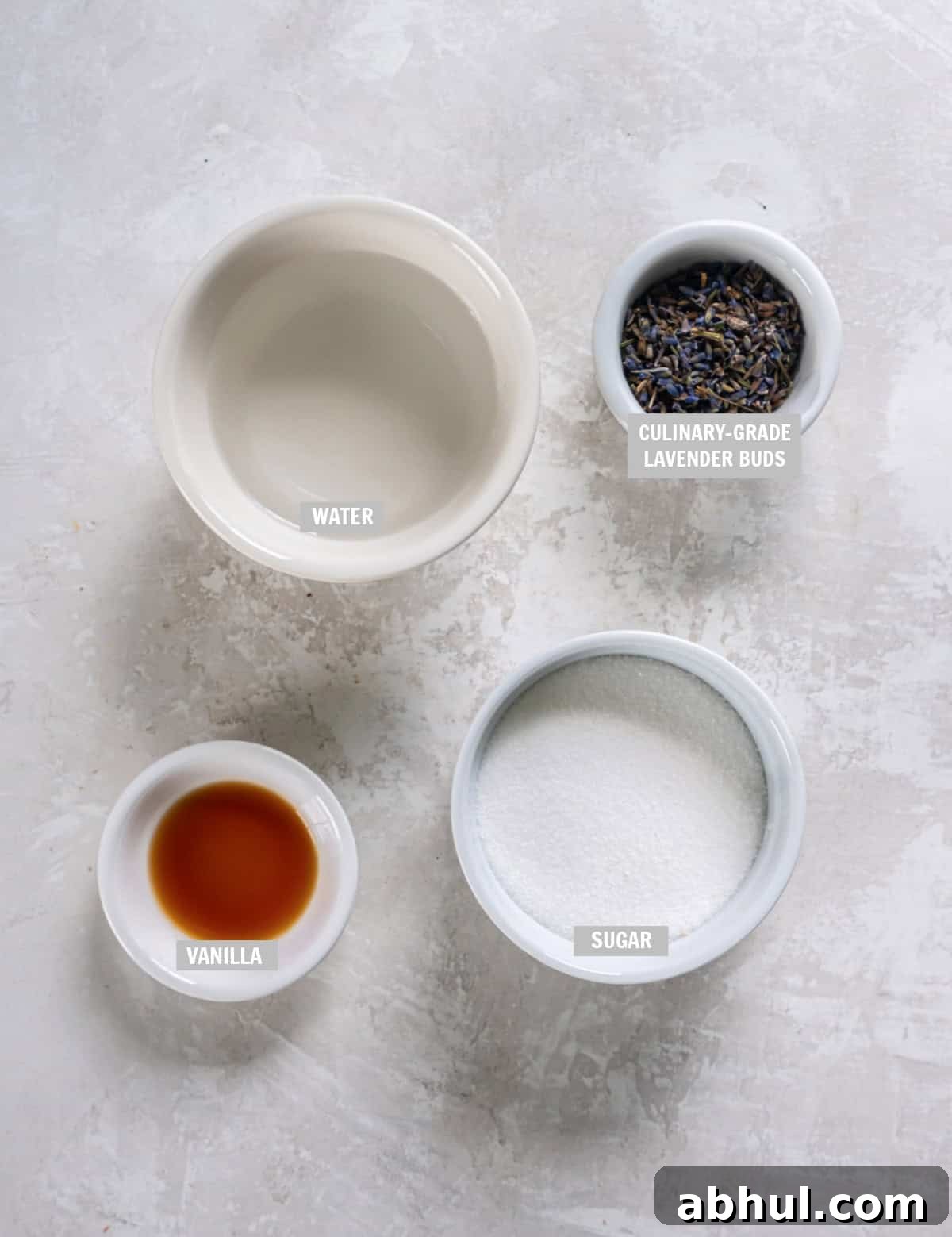 All of the ingredients for lavender simple syrup in bowls on a countertop including sugar, water, lavender buds and vanilla. 