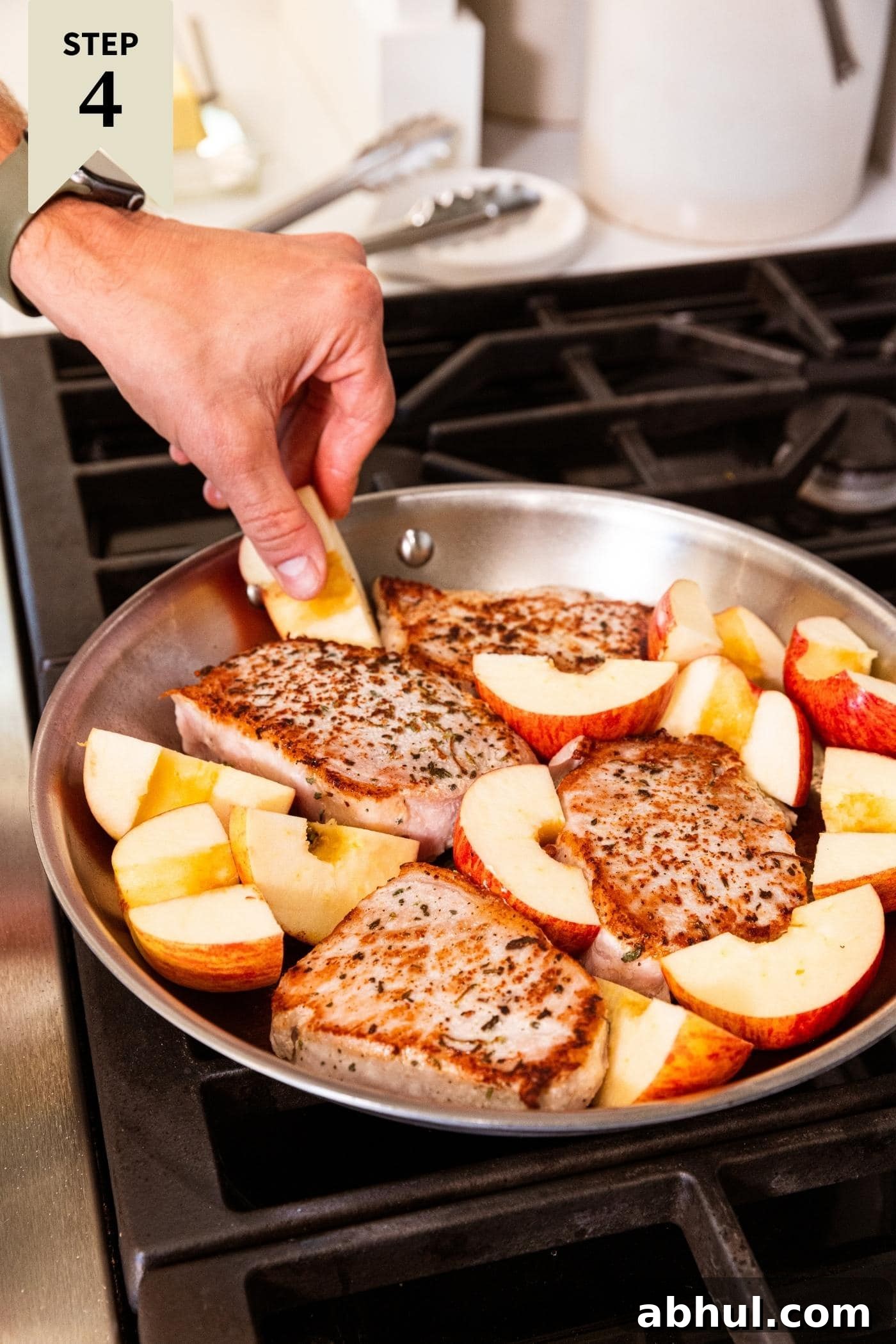 Hand placing apple slices around browned pork chops in metal skillet on stovetop.