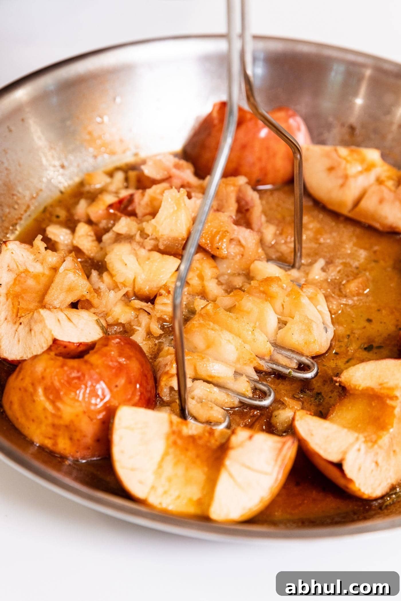 Masher smooshing apples into rough applesauce after being cooked in a skillet in the oven with pan juices collected at the bottom.
