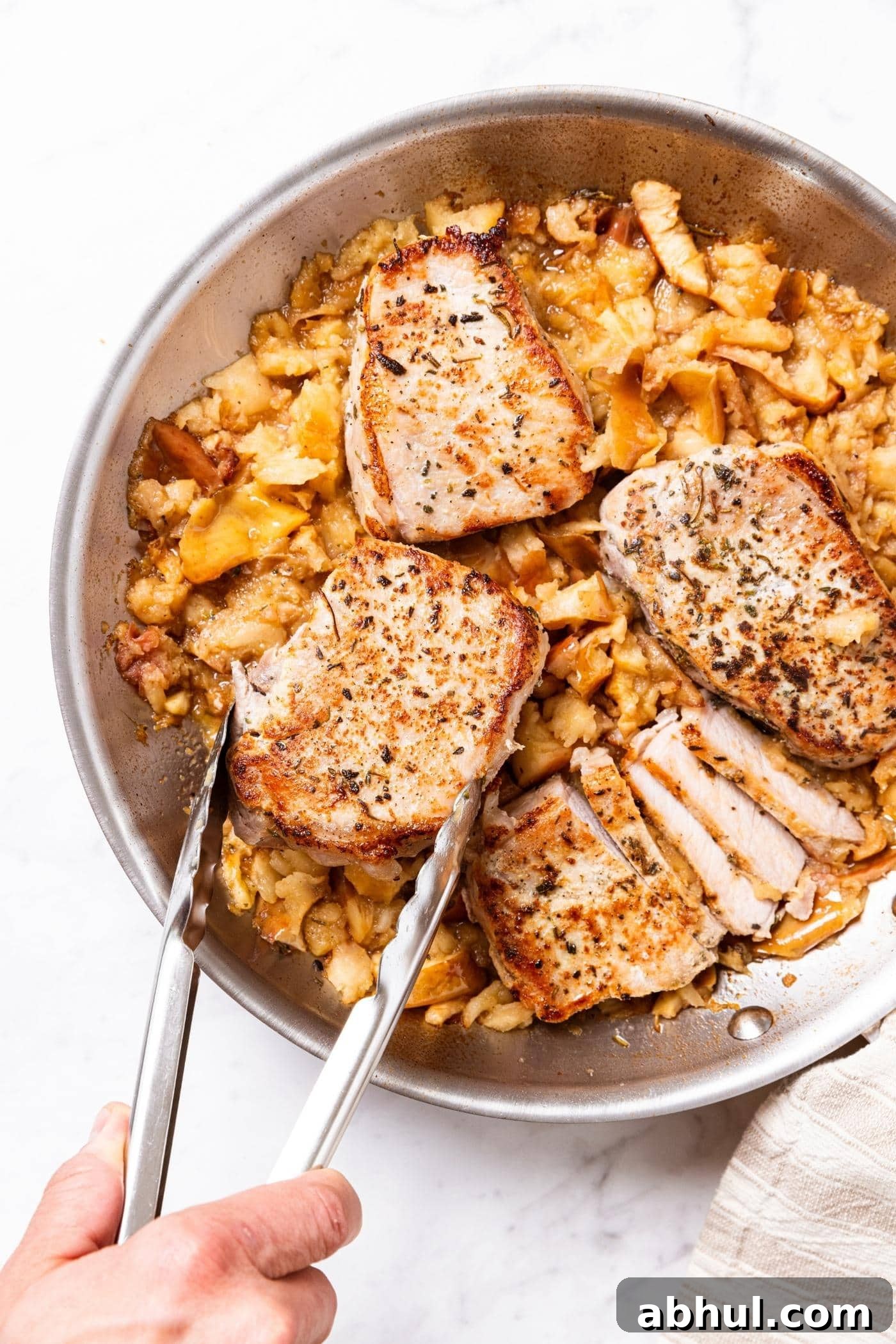 Metal skillet sitting on marble surface filled with smashed apples with browned pork chops being nestled on the top.