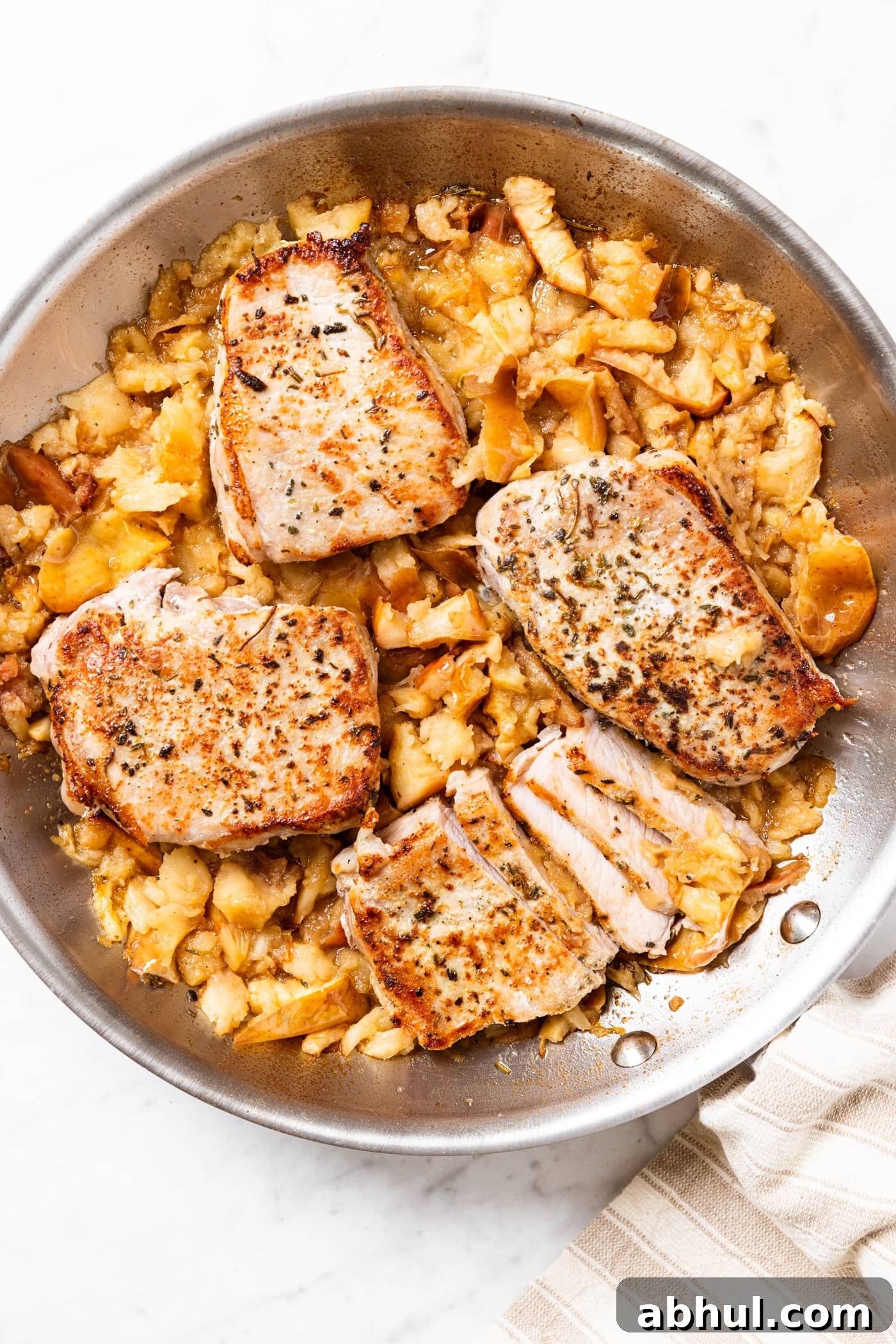 Metal skillet sitting on marble surface filled with smashed apples and four boneless pork chops nestled on top.