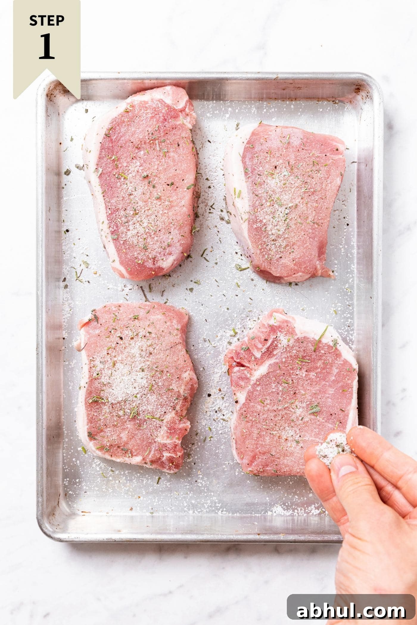 Hand sprinkling dry brine salt mixture on top of raw pork chops sitting on metal pan on marble surface.
