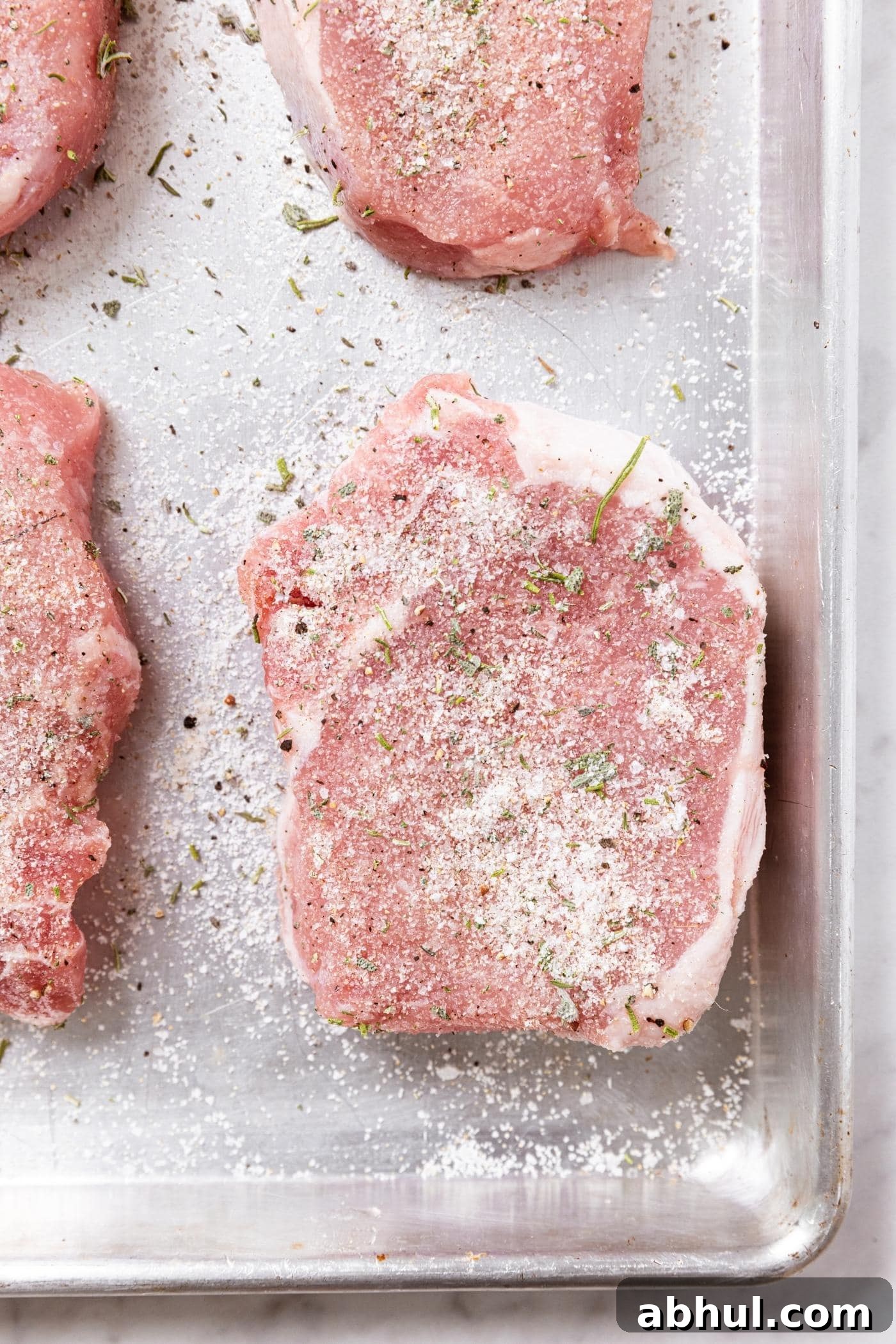 Raw pork chops sitting on metal baking dish with dry brine made from salt sprinkled all over the top.