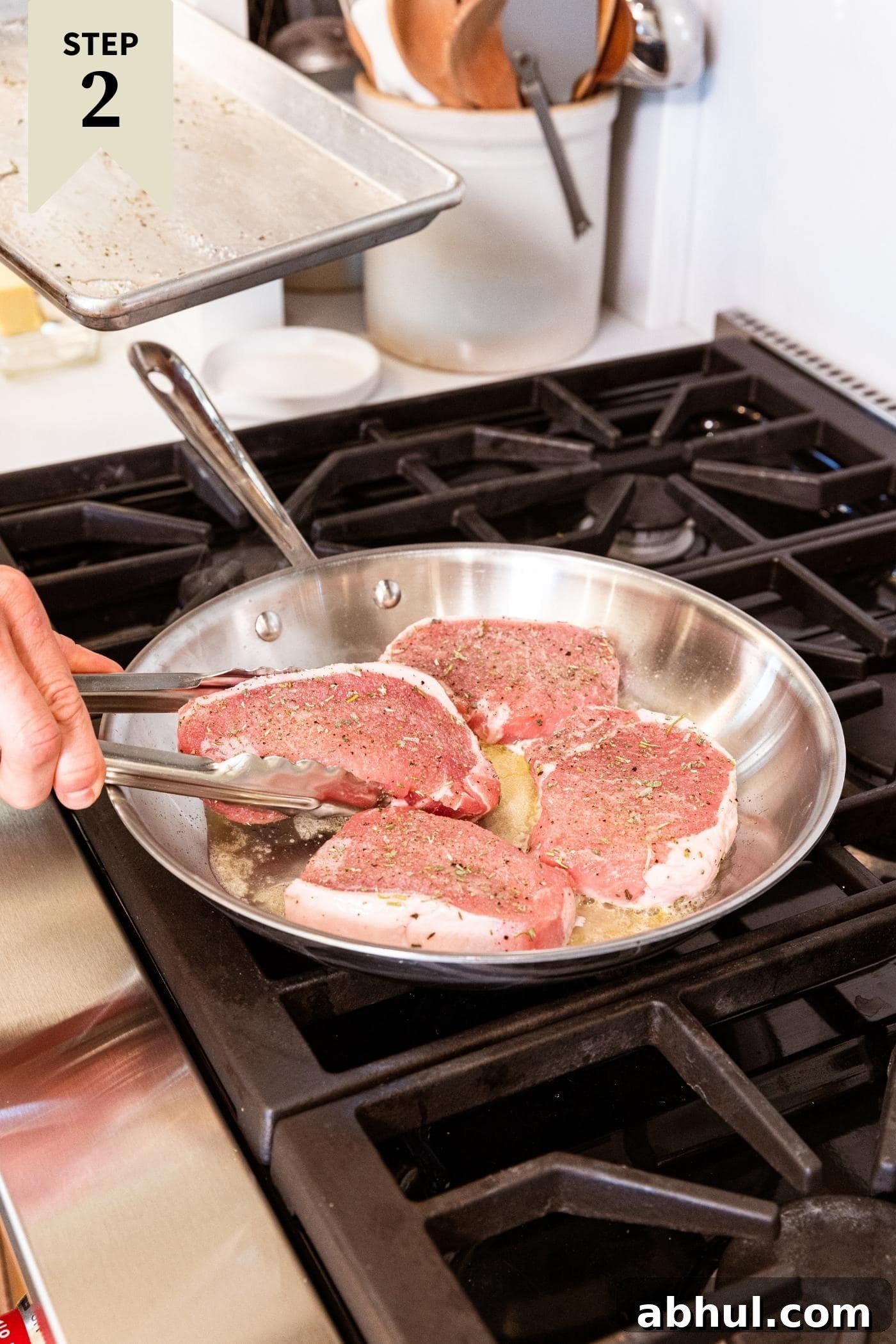 Hand holding tongs browning four pork chops in metal skillet on stovetop.